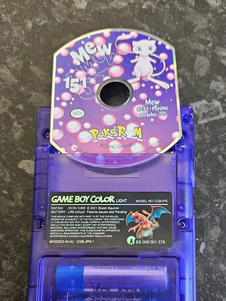 A mini CD labeled "Mew 151 PokéROM PC/Mac Compatible". It is placed slightly into the slot of a clear purple Game Boy Color. A label on the back of the Game Boy shows an image of a Charizard Pokémon and is labled "Game Boy Color Light".