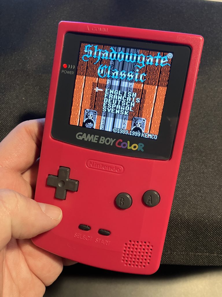 A Game Boy turned on. The title screen for Shadowgate Classic is display. A language selection option is available.