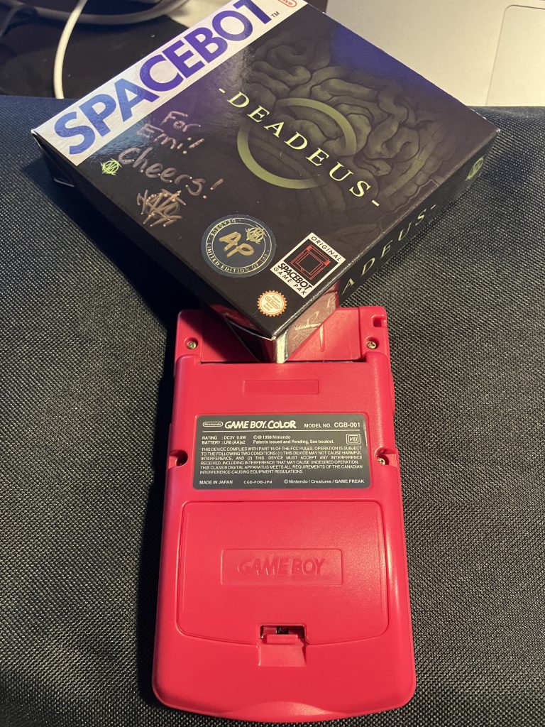 A boxed copy of a Game Boy game attempted to be placed into the empty cartridge slot, which it won't fit in as the box is bigger than the Game Boy. The art on the box displays what looks like an intestine with the game's title, "Deadeus", overlayed. The publisher's name, "Spacebot", is written on the side of the box.