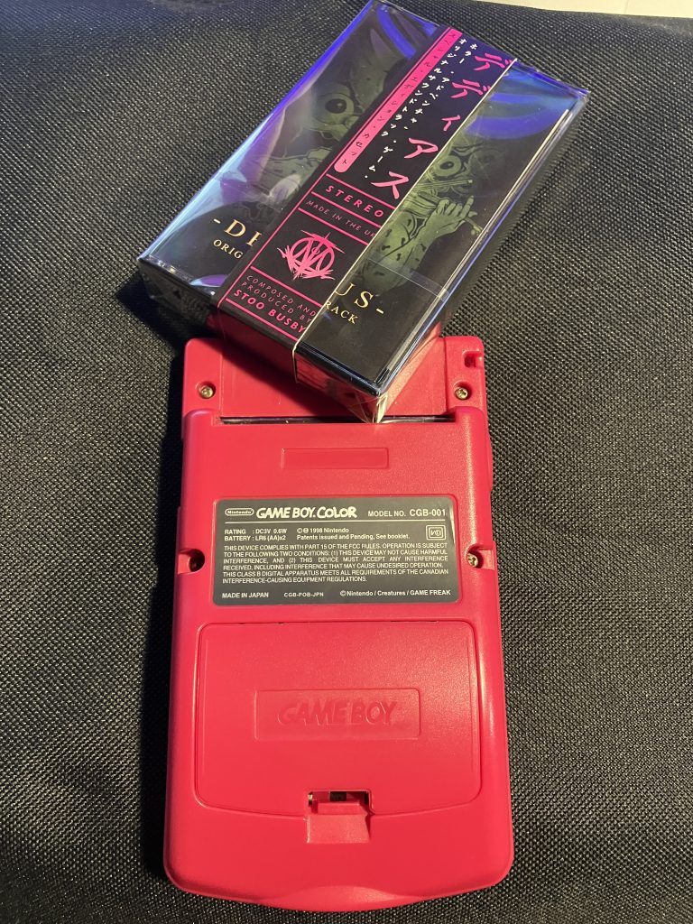 A sealed audio cassette for the soundtrack to the Game Boy Deadeus attempting to be placed in the empty cartridge slot of a Game Boy.